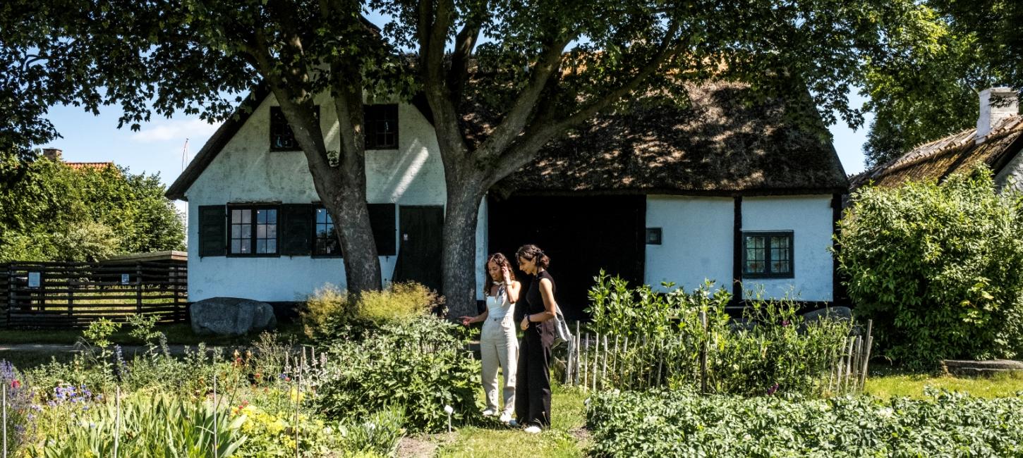 To kvinder i en køkkenhave med gammelt hus i baggrunden