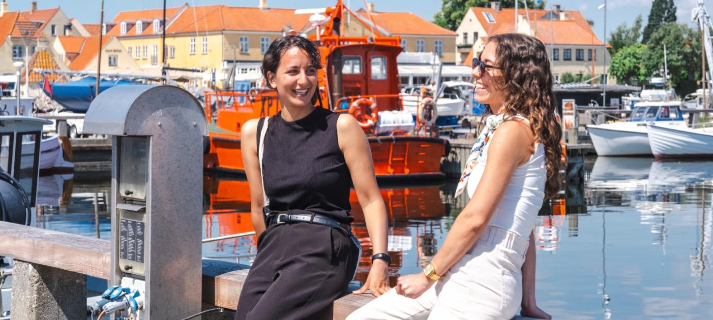 To kvinder sidder på rækværk på havnen