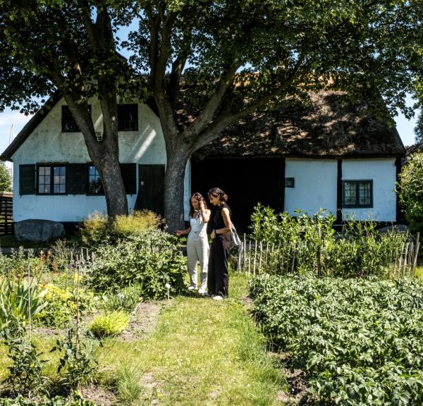 To kvinder i en køkkenhave med gammelt hus i baggrunden